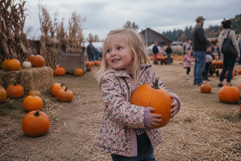 Smiling young girl holding a pumpkin at a rustic pumpkin patch with hay bales and autumn scenery in the background